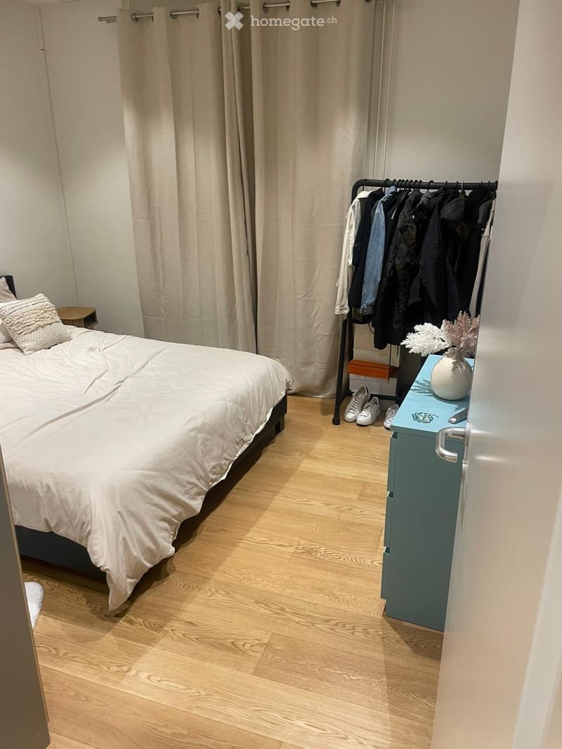 Cozy bedroom with a neatly made bed, a small nightstand, a clothing rack with hanging clothes, and a vase with decorative branches on a blue side table.