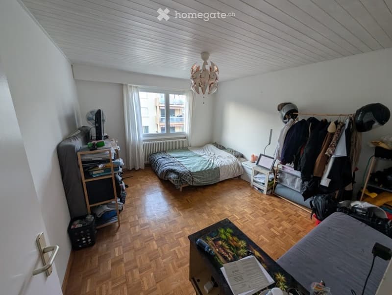 A small, furnished bedroom with wooden flooring, a bed, a table, a chair, a fan, a screen, a radiator, a curtain, a lamp, and a wardrobe with shelves.