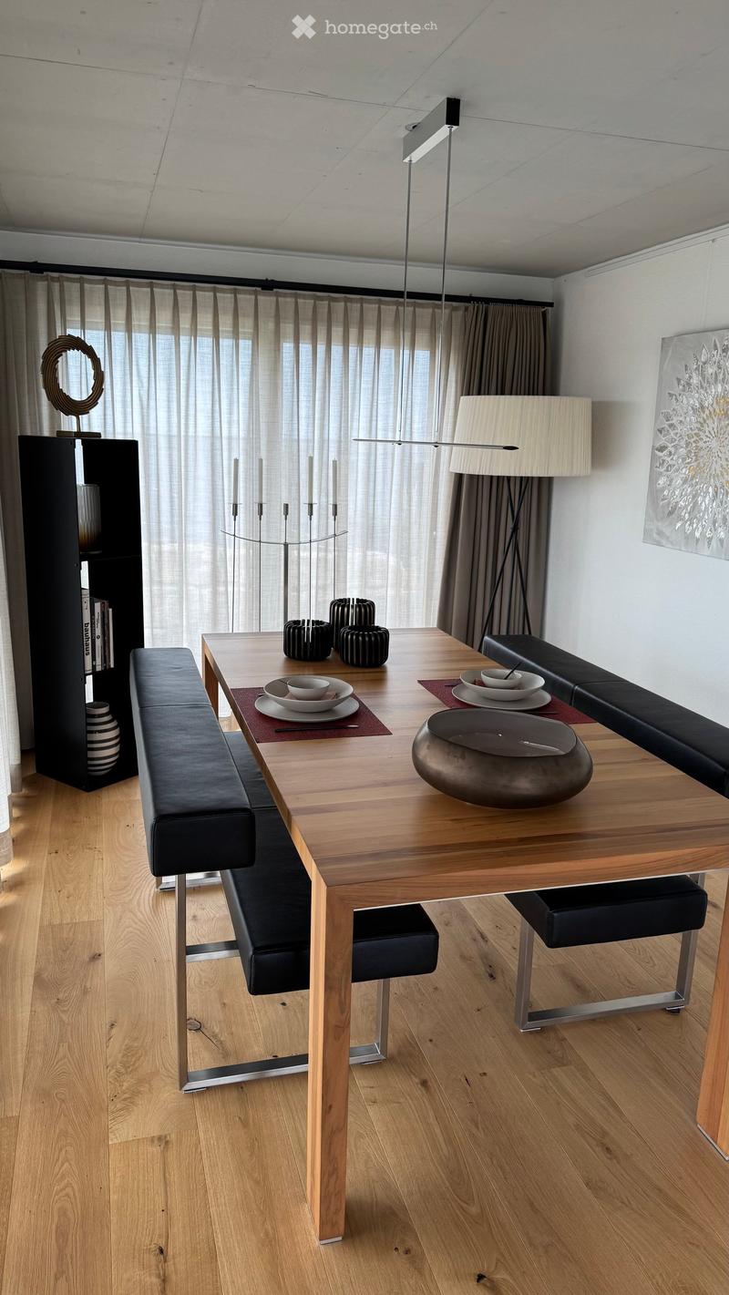 Modern dining area with a wooden table, black leather bench seating, decorative bowls, plates, and a sleek lamp. The room features large windows with sheer curtains and a contemporary aesthetic.