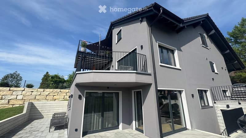 Modern two-story house with grey exterior, multiple balconies, large windows, and a raised ground floor.