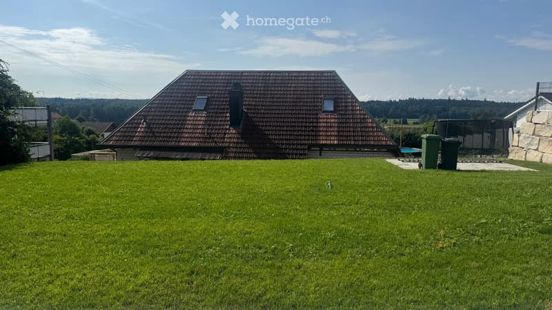 Lush green lawn in front of a house with a tiled roof and multiple windows, surrounded by trees and hills.