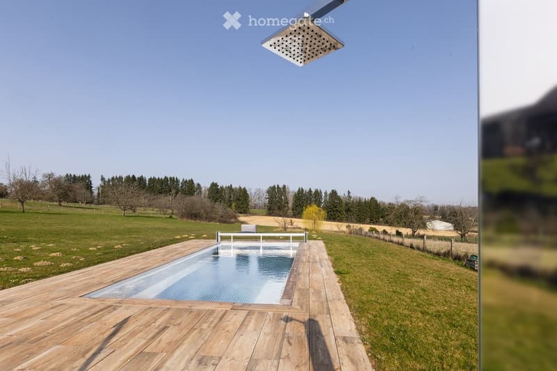 Outdoor area with a swimming pool surrounded by wooden decking, grassy fields, and trees in the background.