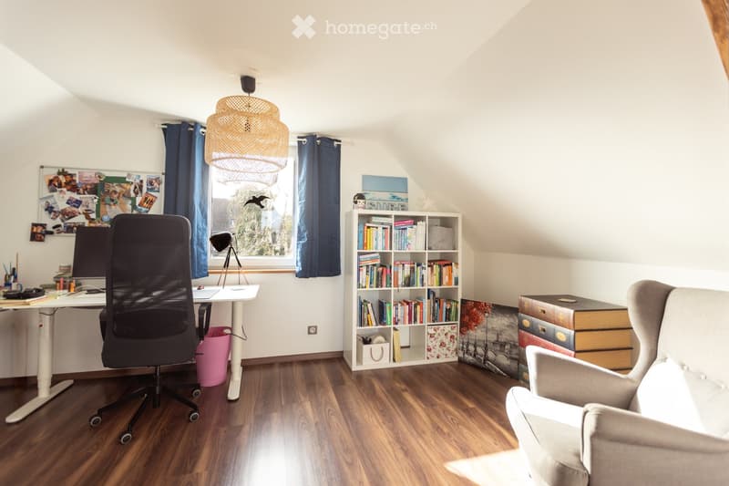 A cozy attic study with wooden flooring, a large window with blue curtains, a modern desk setup, a bookshelf filled with books, and a comfortable armchair.