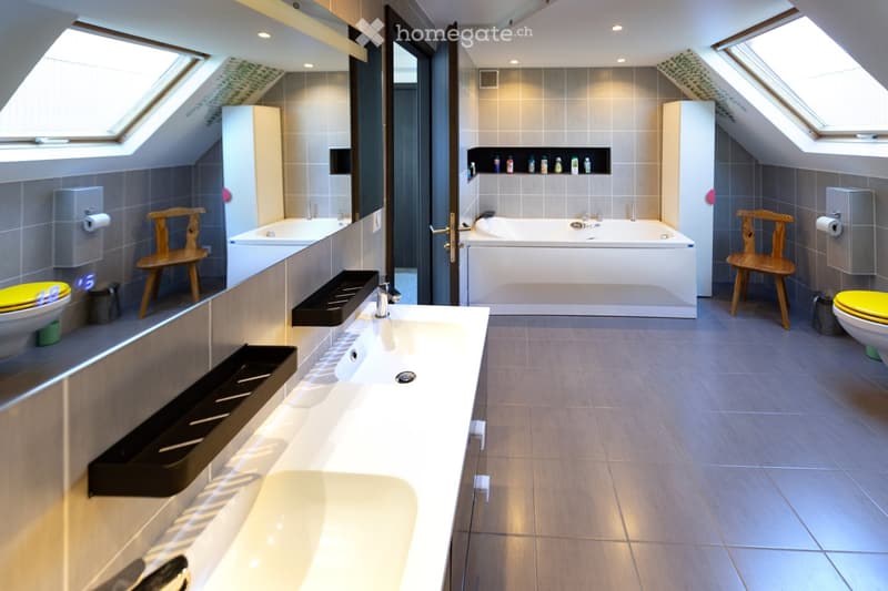Modern bathroom with a double sink, bathtub, toilet, and two wooden chairs. Features include a large mirror, built-in shelves, and skylight windows.