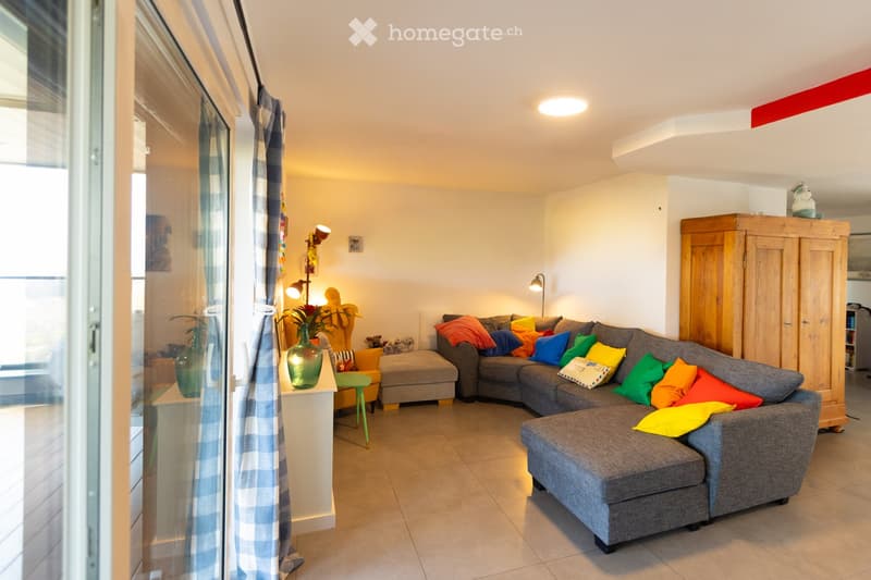 A modern living room with a grey sectional sofa adorned with colorful pillows, a small table with vases, a wooden cabinet, and a green stool. The room features large windows with checkered curtains and warm lighting.