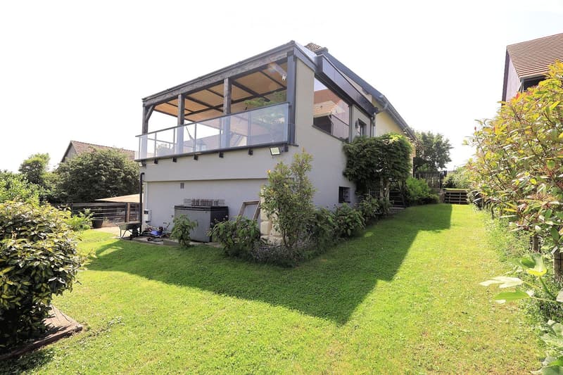 A well-maintained garden with a modern house in the background, featuring a raised terrace with a glass railing, lush greenery, and a neatly trimmed lawn.