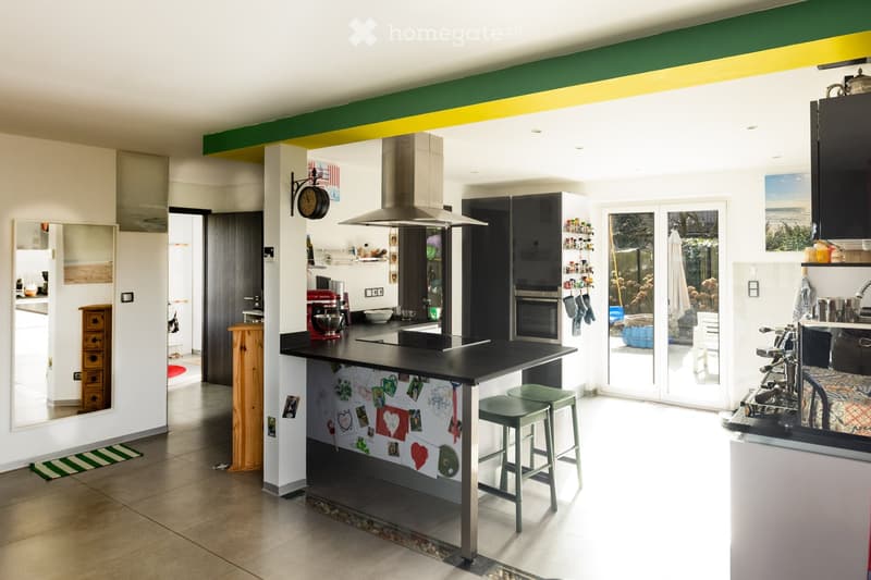 Modern kitchen with a central island, stainless steel appliances, and a black countertop. The island features a backsplash with children's drawings and photos. There are two stools at the island, and the kitchen has a large window with a view of the garde