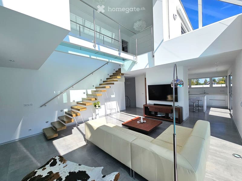 Modern living room with white leather sofa, coffee table, TV stand, and a staircase leading to an upper level. The space is bright with large windows and a sleek design.