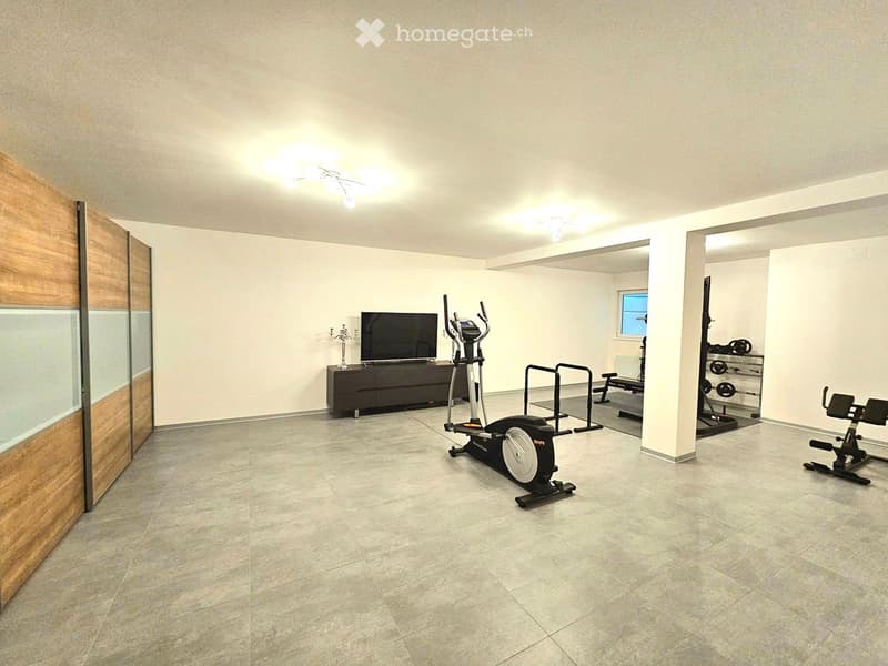 A spacious, well-lit room with tiled flooring and modern lighting. It features a television on a cabinet, an exercise bike, a bench, and various weights, creating a multifunctional space for relaxation and fitness.