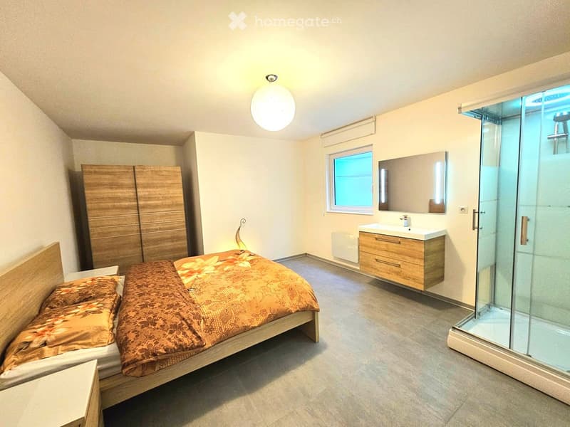 Modern bedroom with a wooden bed, wardrobe, sink, mirror, and glass-enclosed shower. The room features a round ceiling light and a window with blinds.