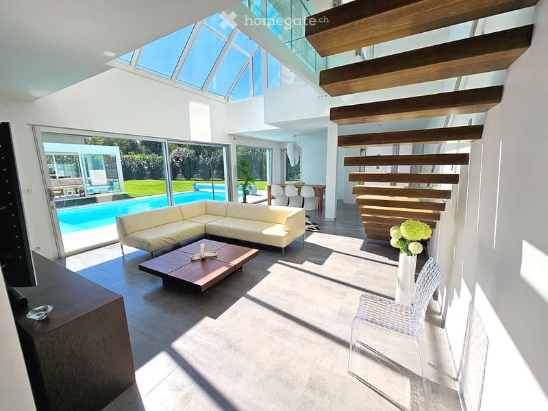Modern living room with large glass windows, a cream-colored sectional sofa, a wooden coffee table, a white vase with flowers, and a clear plastic chair. The room has a high ceiling with wooden beams and a skylight, leading to a pool area outside.