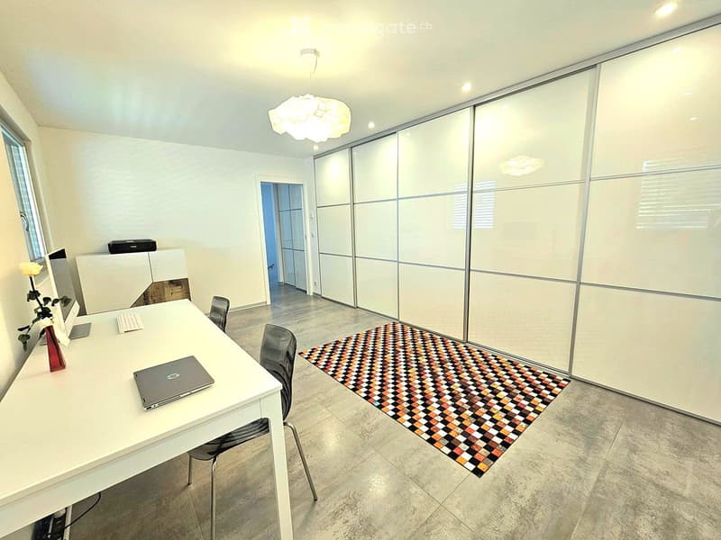 Modern, minimalist room with a white desk, laptop, keyboard, two chairs, a patterned rug, and a large sliding wardrobe. The room features a modern chandelier and a window with blinds.