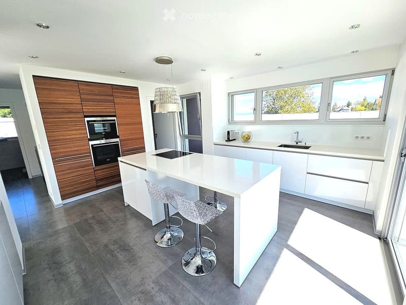 Modern kitchen with white cabinets, a central island, and wooden accents. Features include a built-in oven, sink, and bar stools.