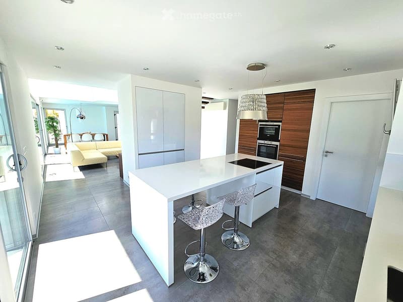 Modern kitchen with white island, built-in appliances, and bar stools. Open plan connects to a living area with a sofa and dining table.