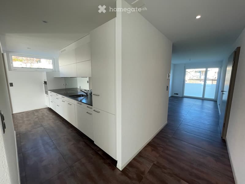Modern, unfurnished kitchen and living area with white cabinets, dark countertops, and large windows. The space features tiled flooring in the kitchen and wooden flooring in the living area, with a sliding glass door leading to a balcony.