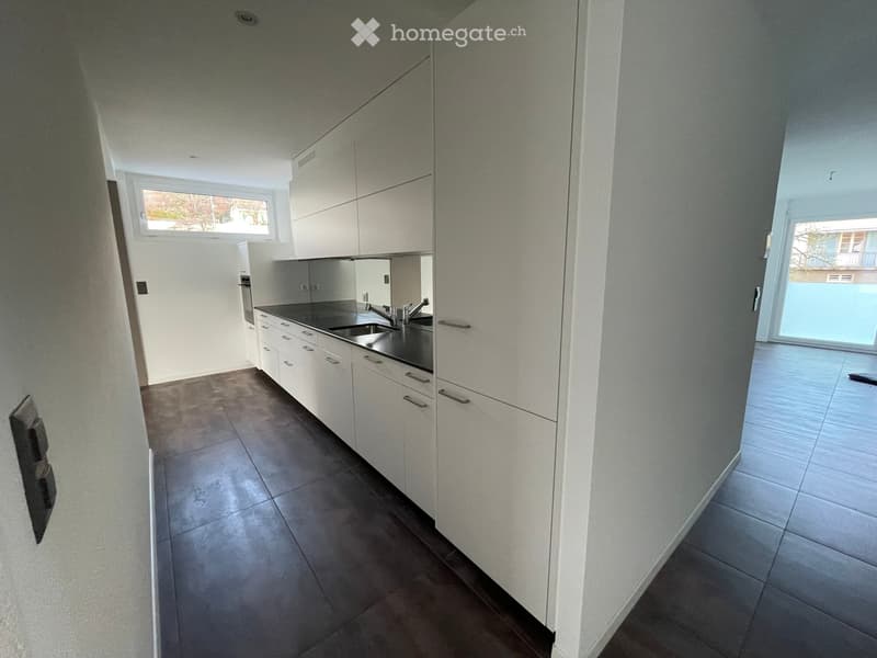 Modern kitchen with white cabinets, dark countertops, and a stainless steel sink. The space is well-lit with natural light from a window and has a tiled floor.