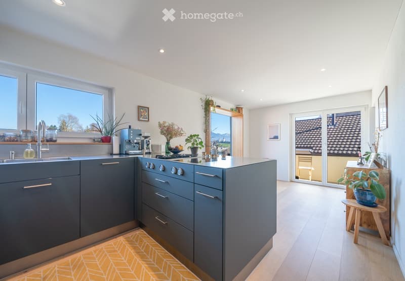 Modern kitchen with grey cabinets, stainless steel appliances, a central island, and a large window overlooking a scenic view. The space includes a sink, coffee maker, stove, and various decorative items such as vases and framed pictures.