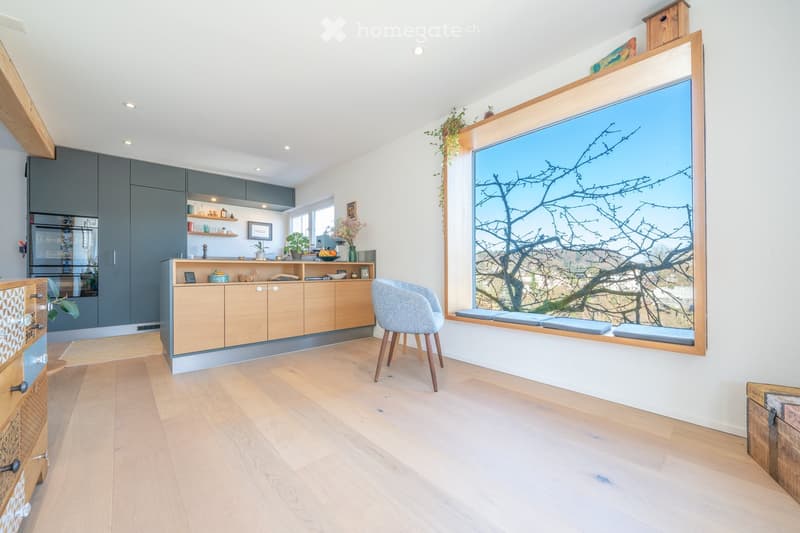 Modern, open-plan space with light wooden flooring, grey and light wood cabinets, a grey chair, and a large window with a scenic view.
