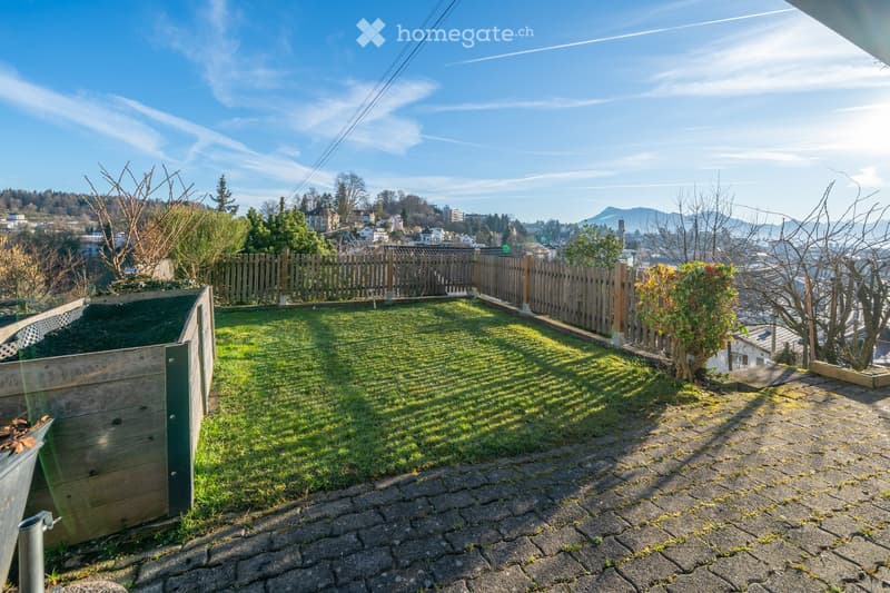 A sunny rooftop garden with a wooden fence, grassy area, and a view of the city and mountains.