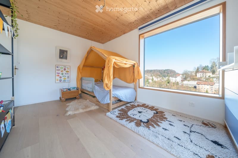 Bright bedroom with wooden floor, large window offering a scenic view, a bed with a yellow canopy, a rug, and a small cabinet with toys.