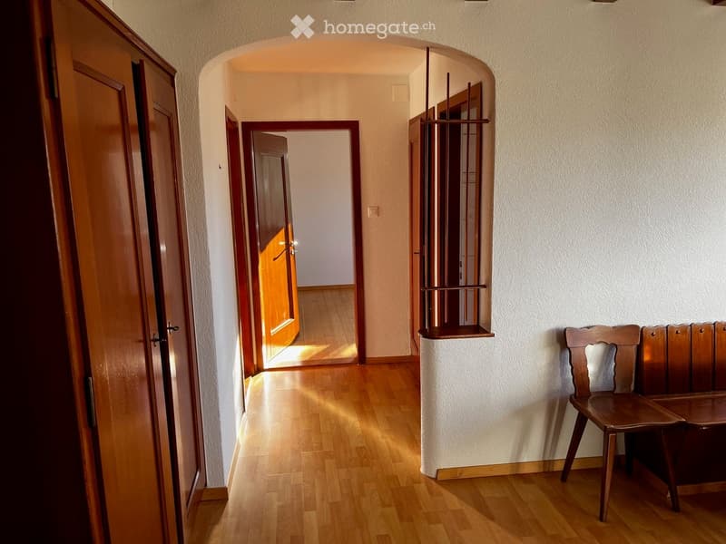 A well-lit hallway with wooden flooring, arched doorways, and a small wooden bench against the wall.