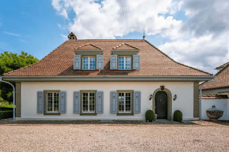 A two-story white house with a red-tiled roof, multiple windows with shutters, and a gravel driveway.