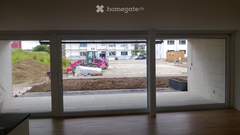 Empty room with large glass sliding doors leading to an outdoor construction area. The interior has a wooden floor and white walls.