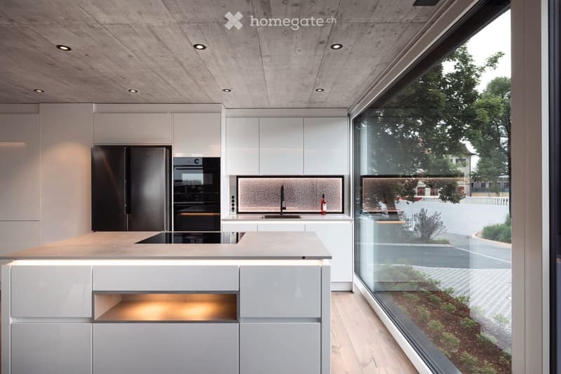 Modern kitchen with white cabinets, a large island, and a sleek black refrigerator. The space features a concrete ceiling with recessed lighting and expansive windows offering a view of the garden.