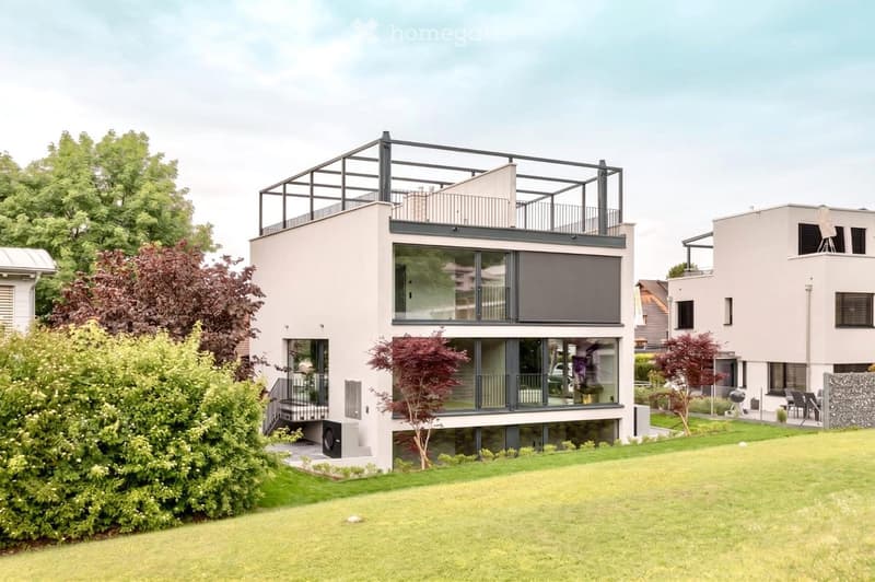 Modern two-story house with large glass windows, a raised ground floor, and a rooftop terrace with a metal railing.