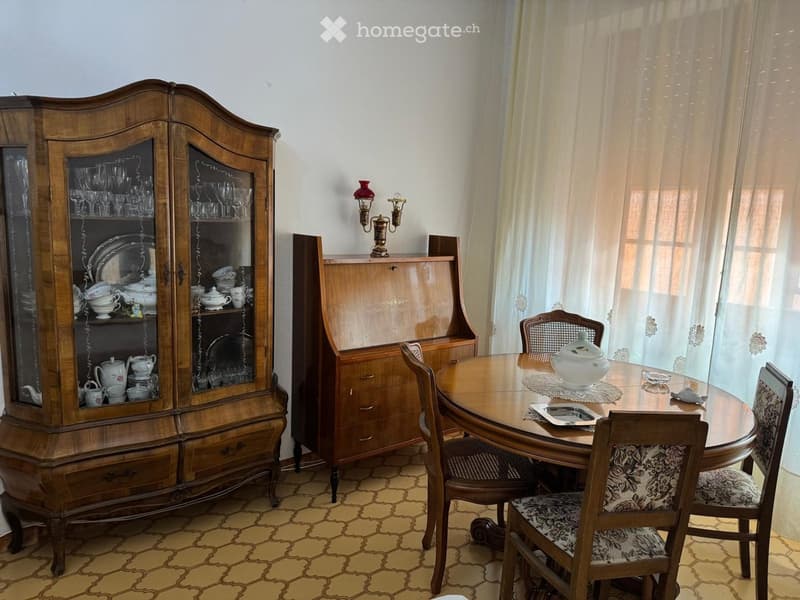 Dining room with a round wooden table, six chairs, a large wooden cabinet with glass doors, and decorative items on the table and cabinet.