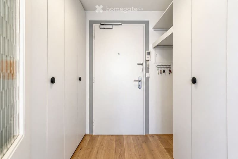 A modern, minimalist hallway with light wooden flooring, white walls, and a closed door leading to another room. A small built-in shelf unit with hooks for keys is visible on the right side.