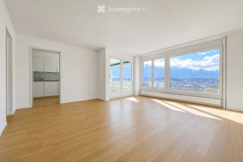 Empty room with wooden flooring, white walls, large windows offering a scenic mountain view, and a small kitchen area with cabinets and a sink.