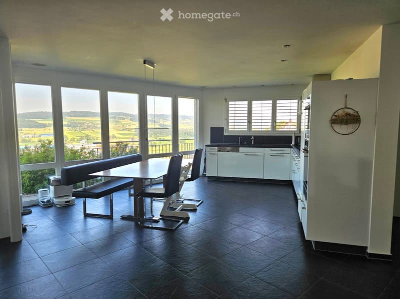 Modern kitchen and dining area with large windows offering a scenic view, featuring a dark tiled floor, a long dining table with bench seating, and sleek white kitchen cabinets.