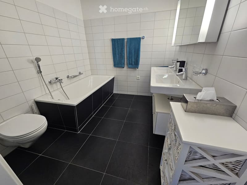 Modern bathroom with white tiles, black floor tiles, bathtub, toilet, double sink, mirror, and two blue towels hanging on a rack.