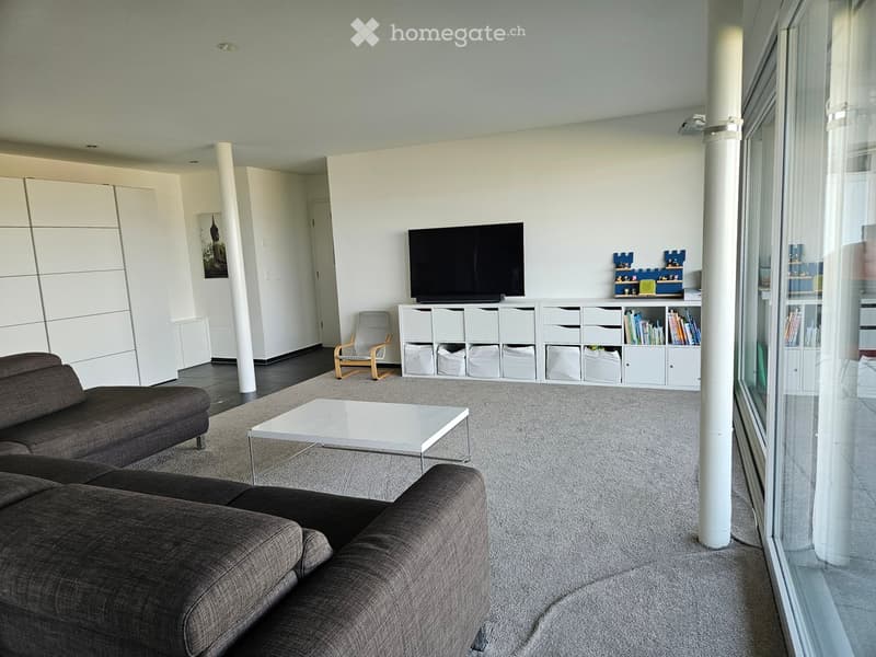 Modern living room with a grey sectional sofa, white coffee table, wooden chair, TV on a white storage unit, and large windows.