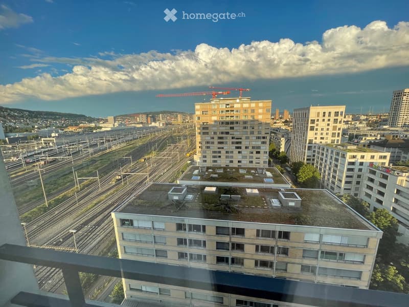 View from a balcony showing a cityscape with multiple residential buildings, train tracks, and a partly cloudy sky.