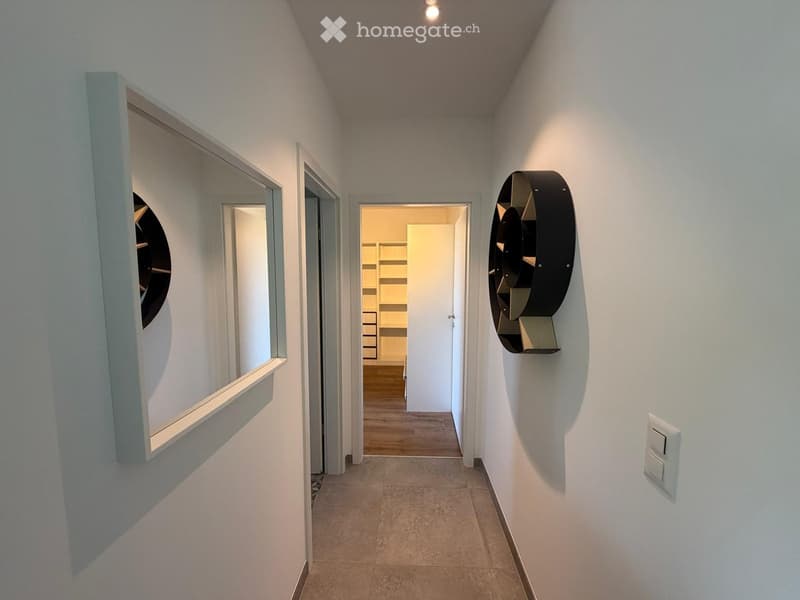 Modern hallway with white walls, tiled floor, and recessed lighting. Features a large mirror on the left and a decorative wall-mounted sculpture on the right, leading to an open doorway.