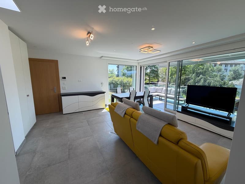 Modern living room with large windows offering a view, a yellow sofa, a coffee table, a television, and a dining area with a table and chairs.