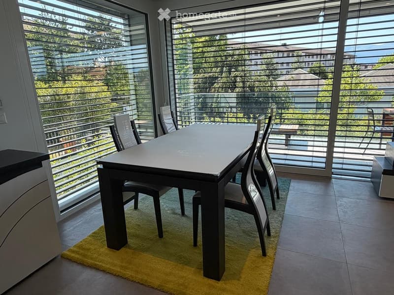 A modern dining area with a sleek black dining table and matching chairs, placed on a yellow rug. Large windows with blinds offer a scenic view of the surrounding greenery and buildings.