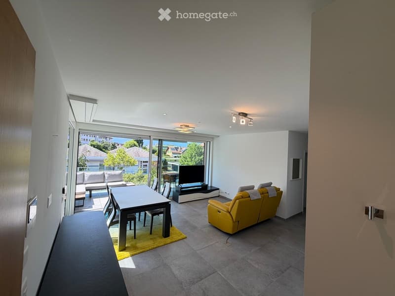 Modern living room with a yellow sofa, dining table, chairs, and a television. Large windows offer a view of the outdoor area.