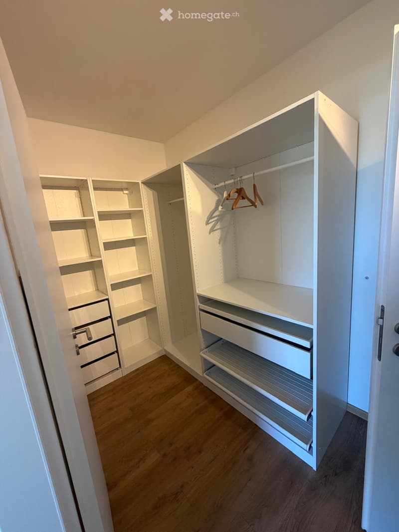 Empty walk-in closet with white built-in shelving units, drawers, and hanging rods. Wooden flooring and white walls.