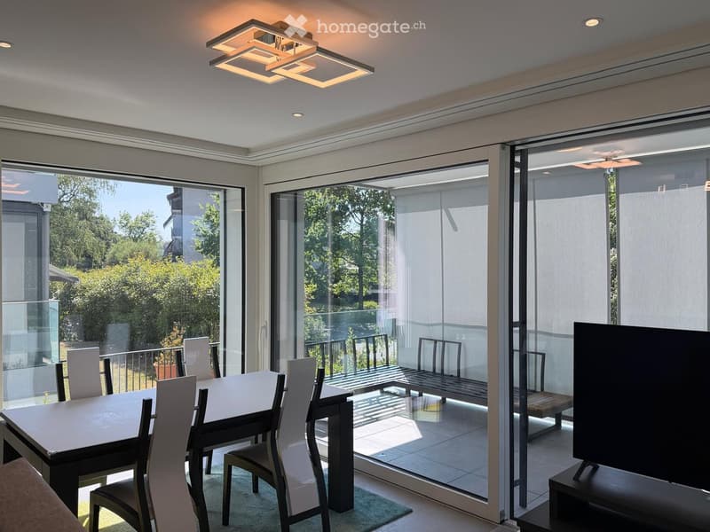 Modern dining area with a sleek white dining table and chairs, large glass sliding doors leading to a balcony, and a television on a stand.