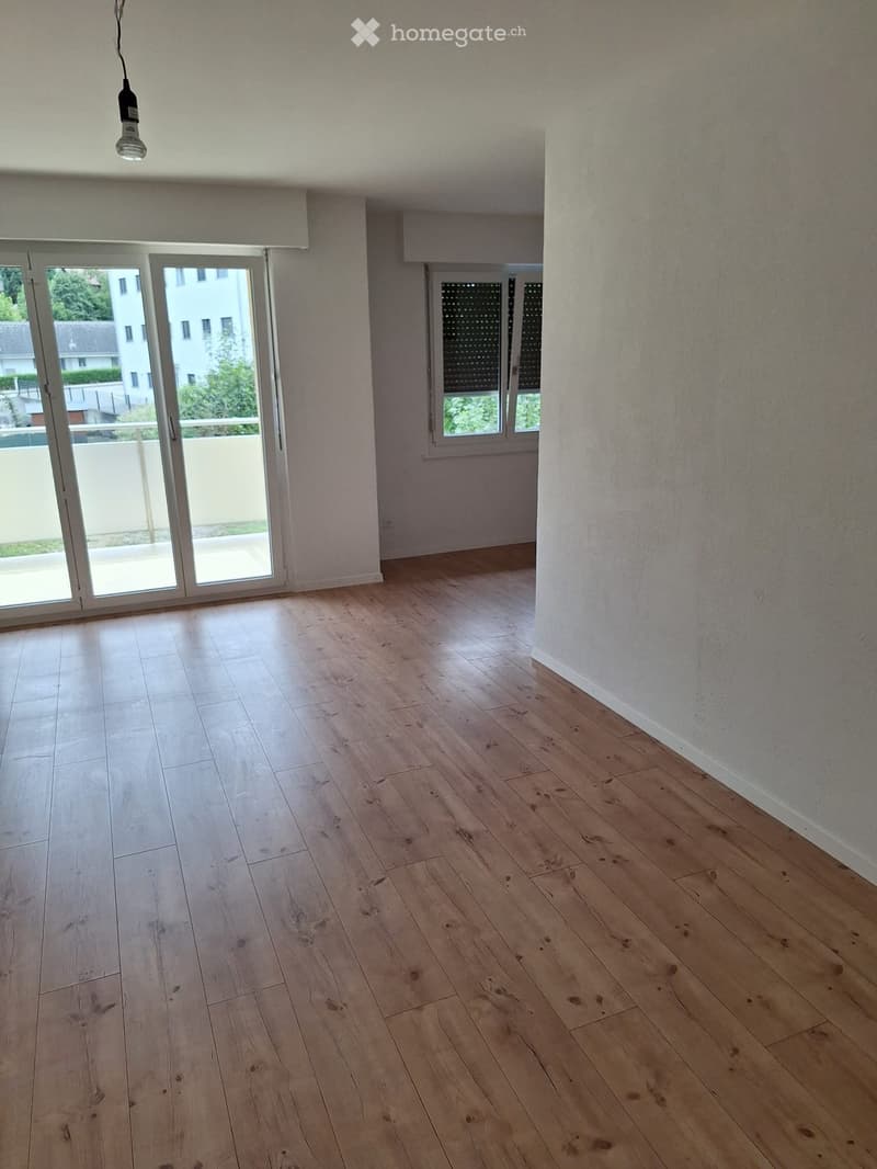 Empty room with light wooden flooring, white walls, large sliding glass door leading to a balcony, and a window with blinds.