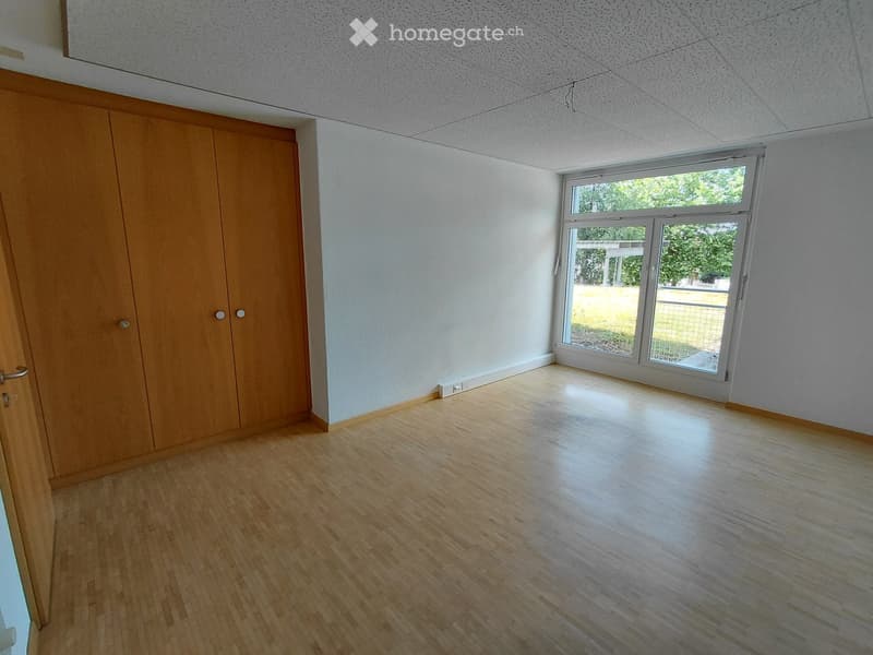 Empty room with wooden flooring, white walls, and a large window with a view of greenery outside. A wooden closet is visible on the left side.