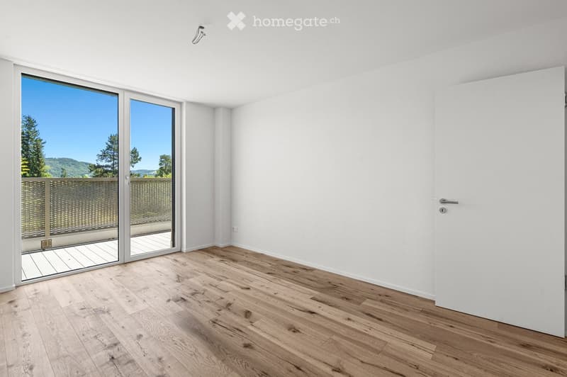 Empty room with light wooden flooring, white walls, large sliding glass doors leading to a balcony, and a view of trees and hills.