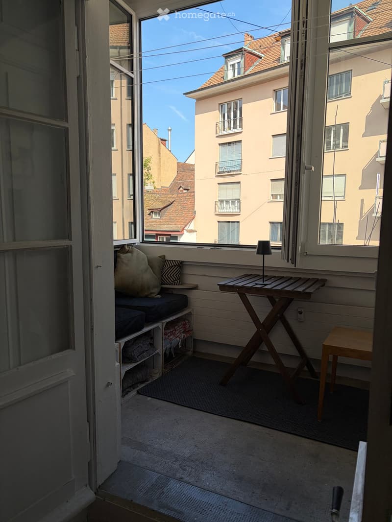 Balcony with wooden table, stool, bench, and lamp, overlooking a cityscape with buildings and clear sky.