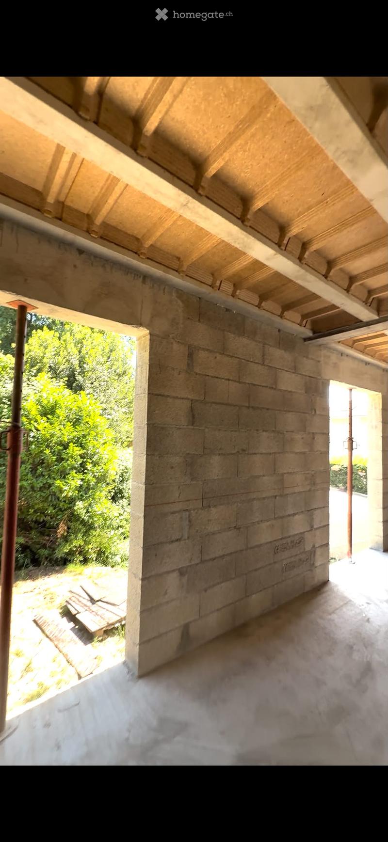 An open, unfinished corridor with exposed wooden beams and stone walls, leading to a garden area with greenery visible through an opening.