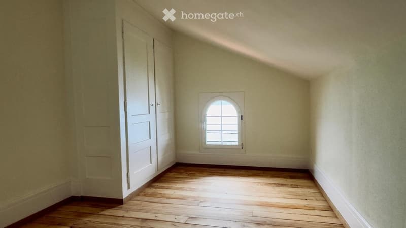 Empty room with wooden flooring, white walls, and a single arched window with multiple panes. There is a white door on the left side.