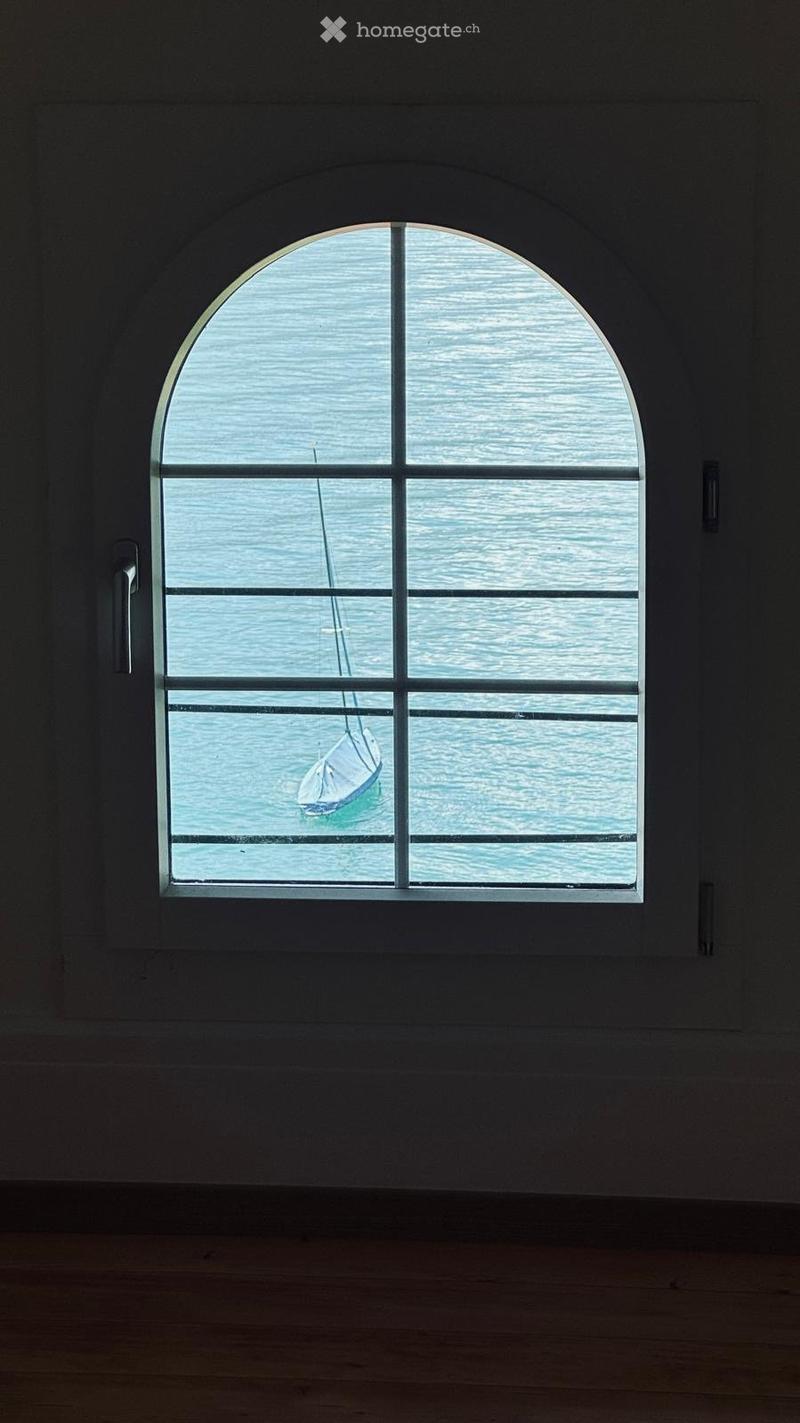 Empty room with wooden floor and large arched window showing a sailboat on the water outside.