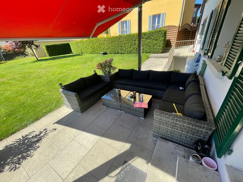 Balcony with a black wicker sofa and coffee table, overlooking a well-maintained lawn and garden.
